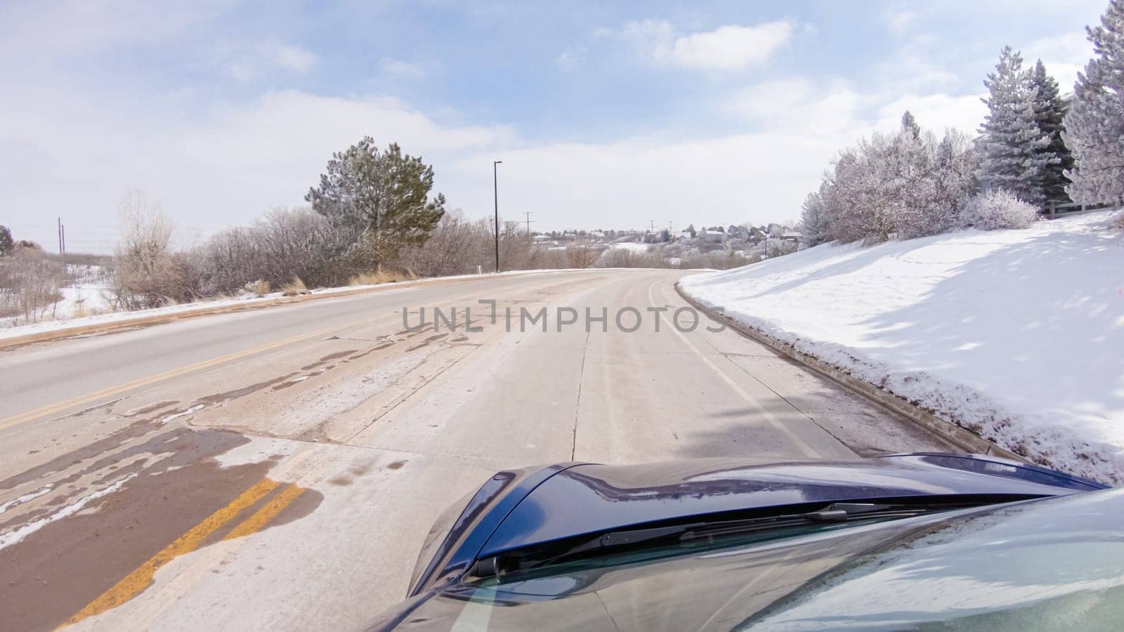Traversing a freshly cleared, suburban road after a winter storm, one experiences a serene drive through an upscale residential neighborhood. Snow-covered houses and trees contribute to a picturesque winter scene, enhancing the tranquility of the journey.