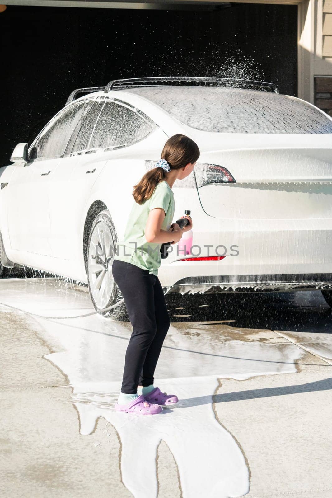 A young girl enthusiastically assists in washing the family's electric car in their suburban driveway.