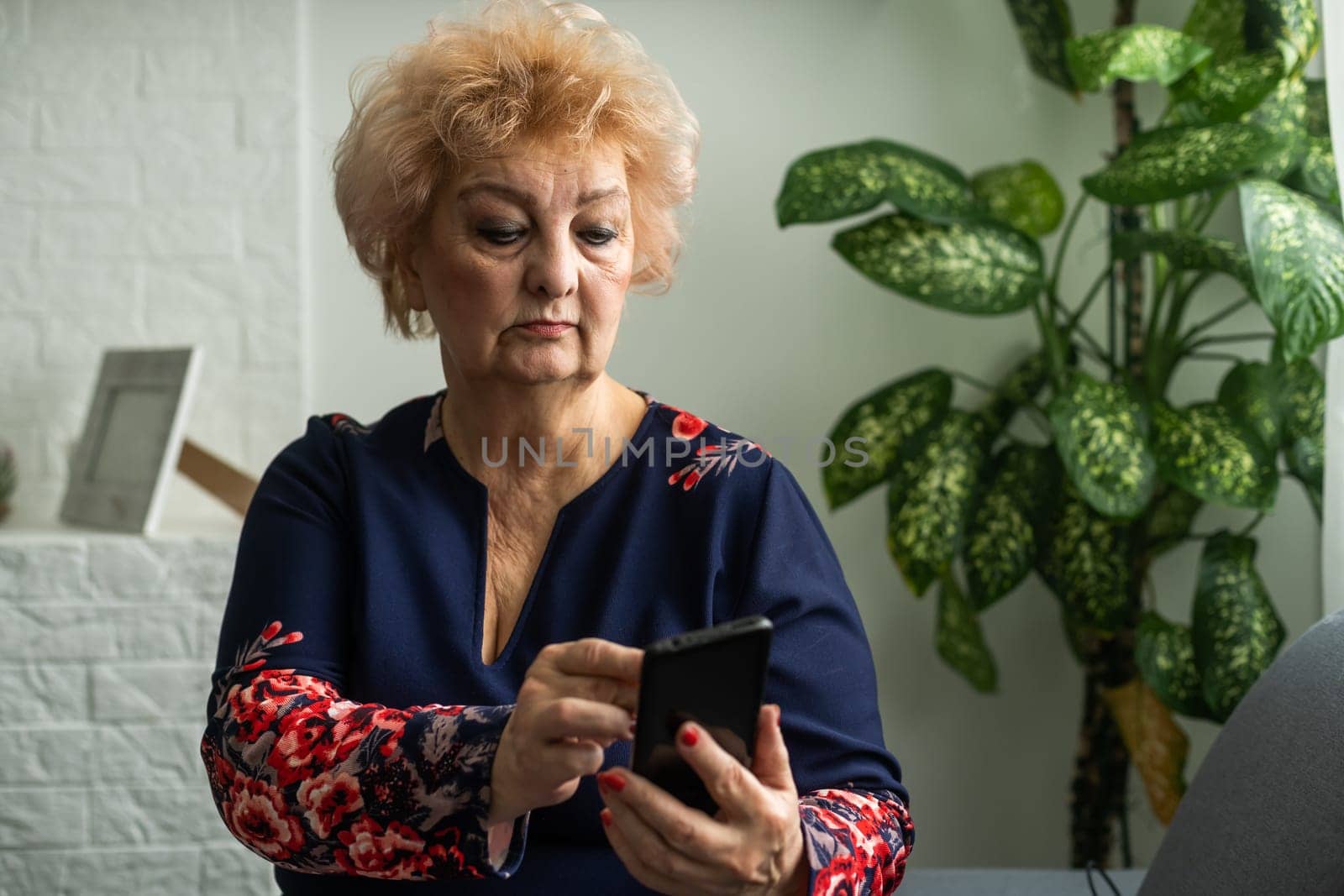 Relaxed mature old 60s woman, older middle aged female customer holding smartphone using mobile app, texting message, search ecommerce offers on cell phone technology device sitting on couch at home.