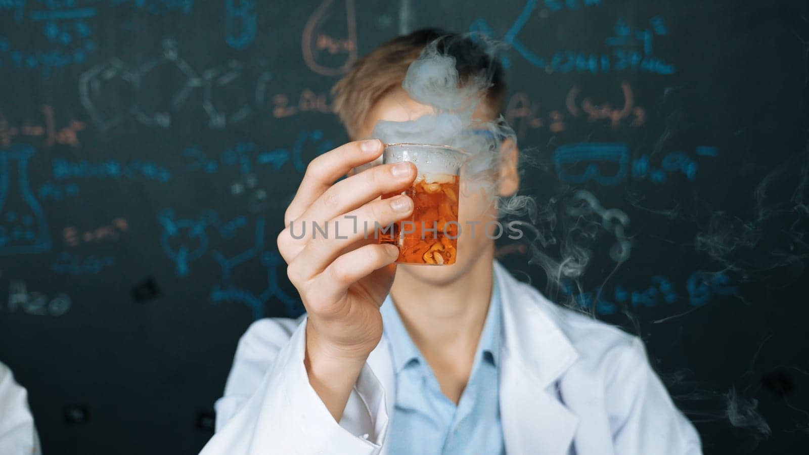 Closeup of boy inspect chemical solution while holding beaker at blackboard with chemical theory. Smart scientist doing experiment while analysis colored solution while wearing lab coat. Edification.