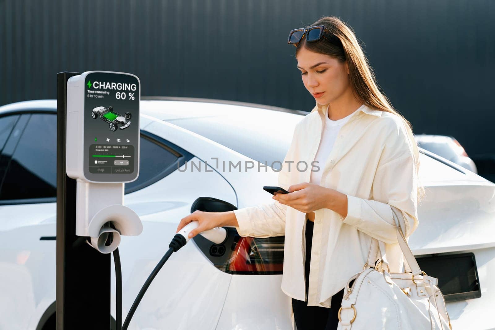 Young woman holding shopping bag and use smartphone to pay for electricity for recharging EV car battery from charging station at city mall parking lot. Modern woman go shopping by eco car. Expedient