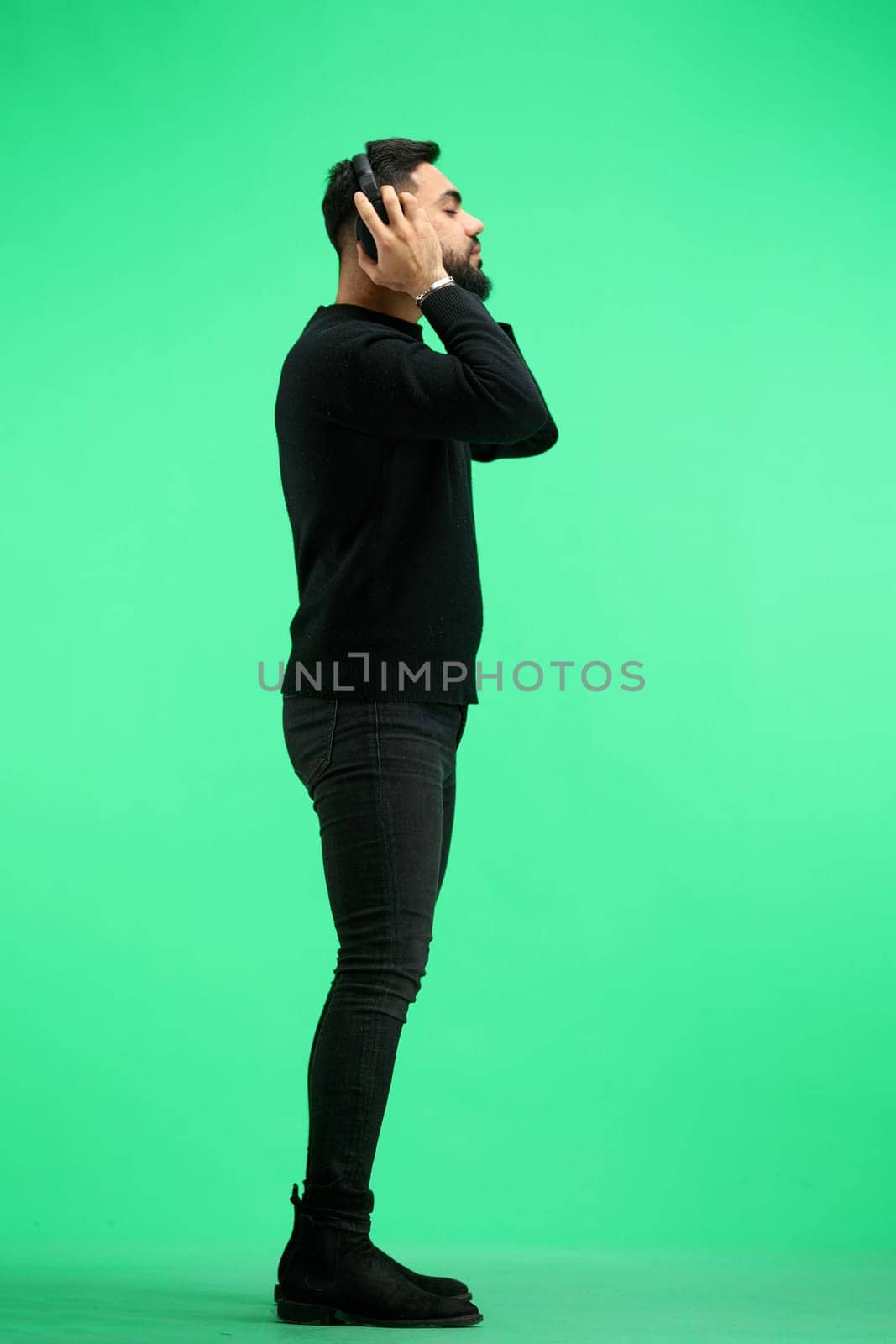 A man, in full height, on a green background, listens to music with headphones.