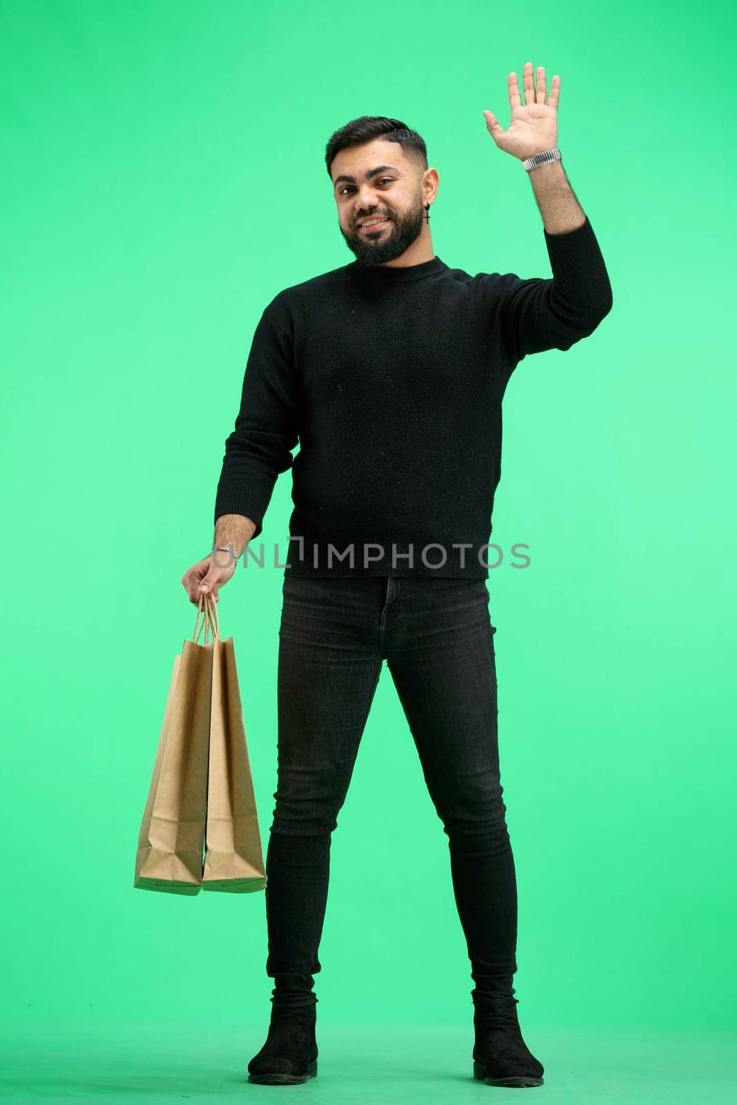 A man, full-length, on a green background, with bags, waving his hand.