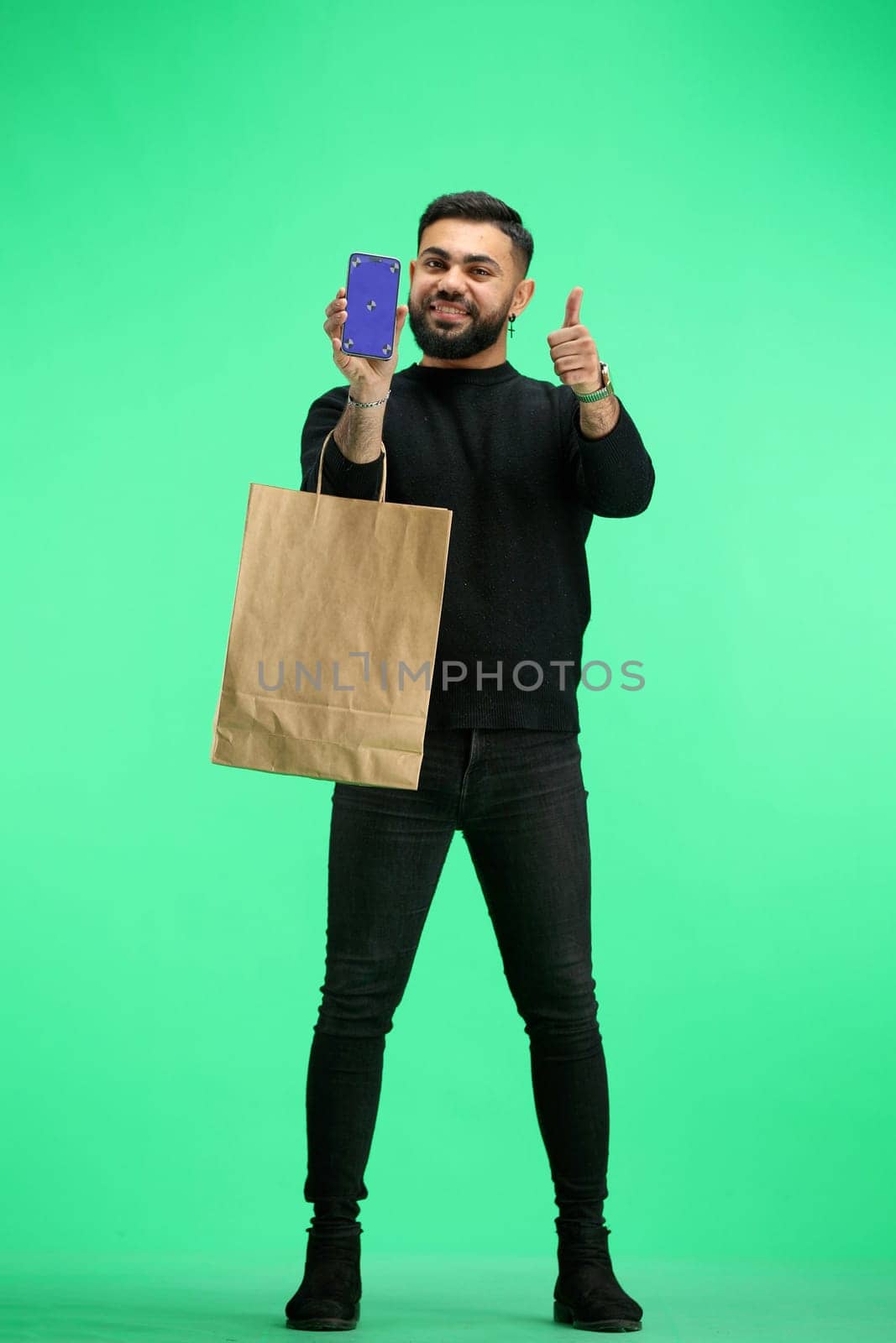 A man, full-length, on a green background, with bags, shows a phone.