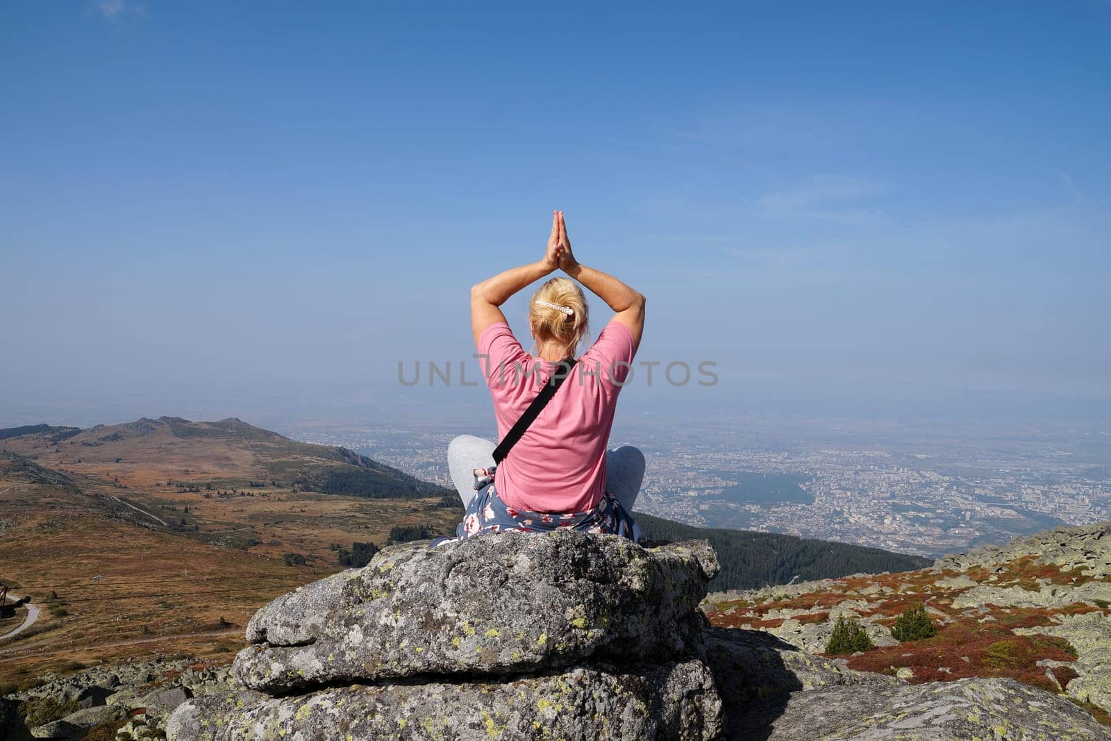 blonde woman meditates in a yoga pose with her arms raised above her head on the top of a mountain, back view