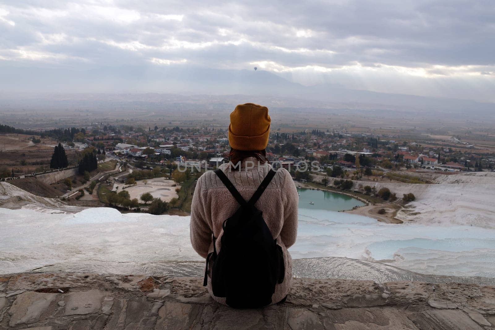 girl with a backpack in a yellow hat sits on the top of Pamukkale, Tyrkey, solo travel, rear view.