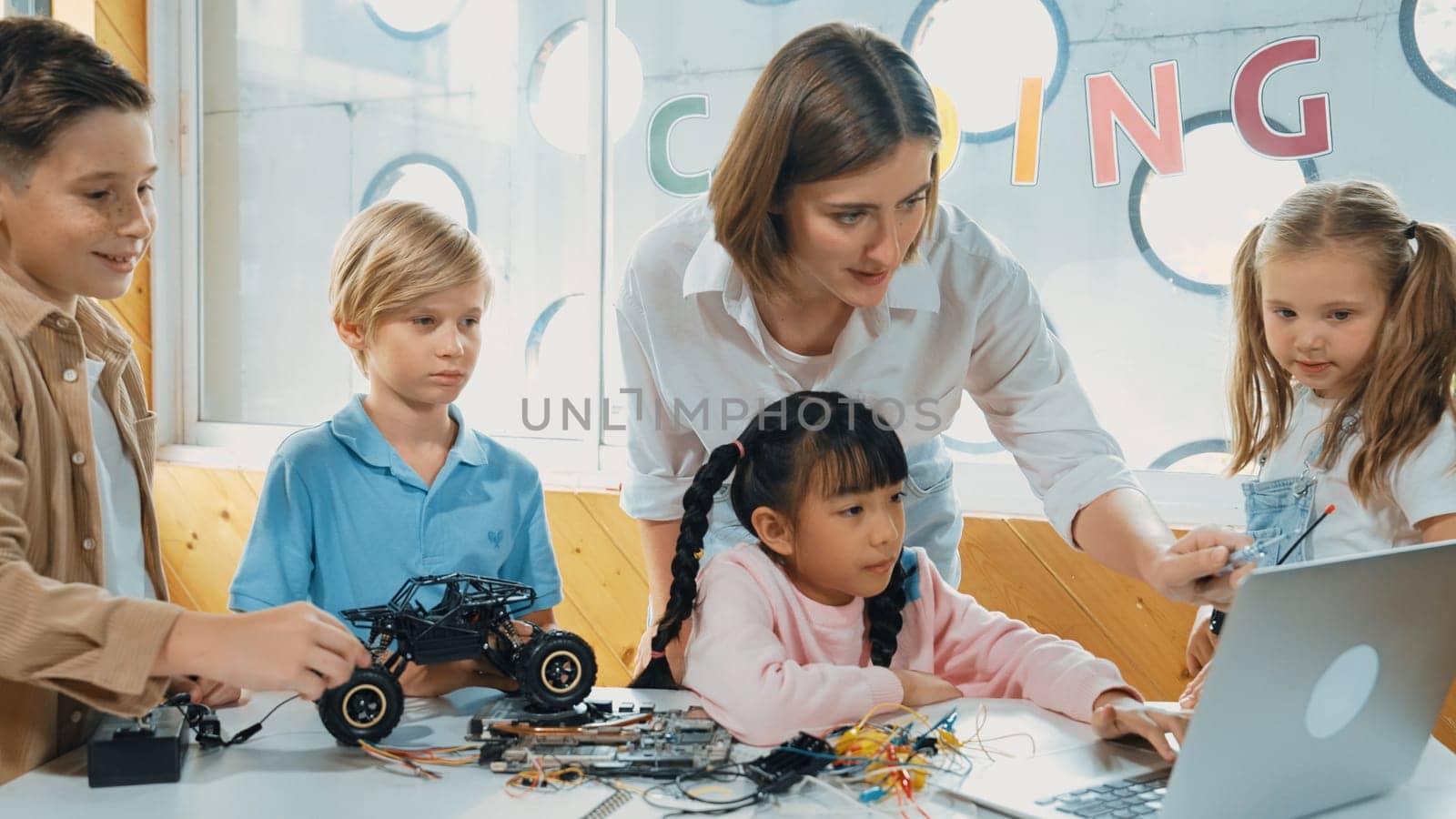 Smart teacher and diverse student looking at laptop on table with car model placed. Caucasian mentor inspect, explain, teaching engineering code or prompt in STEM technology classroom. Erudition.