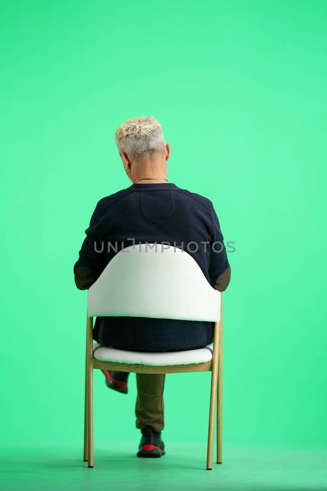 A man, full-length, on a green background, sitting on a chair.