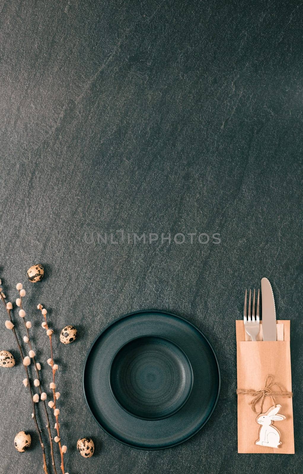 Empty dark plates with a knife, a fork in a cardboard napkin and willow branches with easter eggs lie on a black stone table with copy space from above, flat lay close-up.