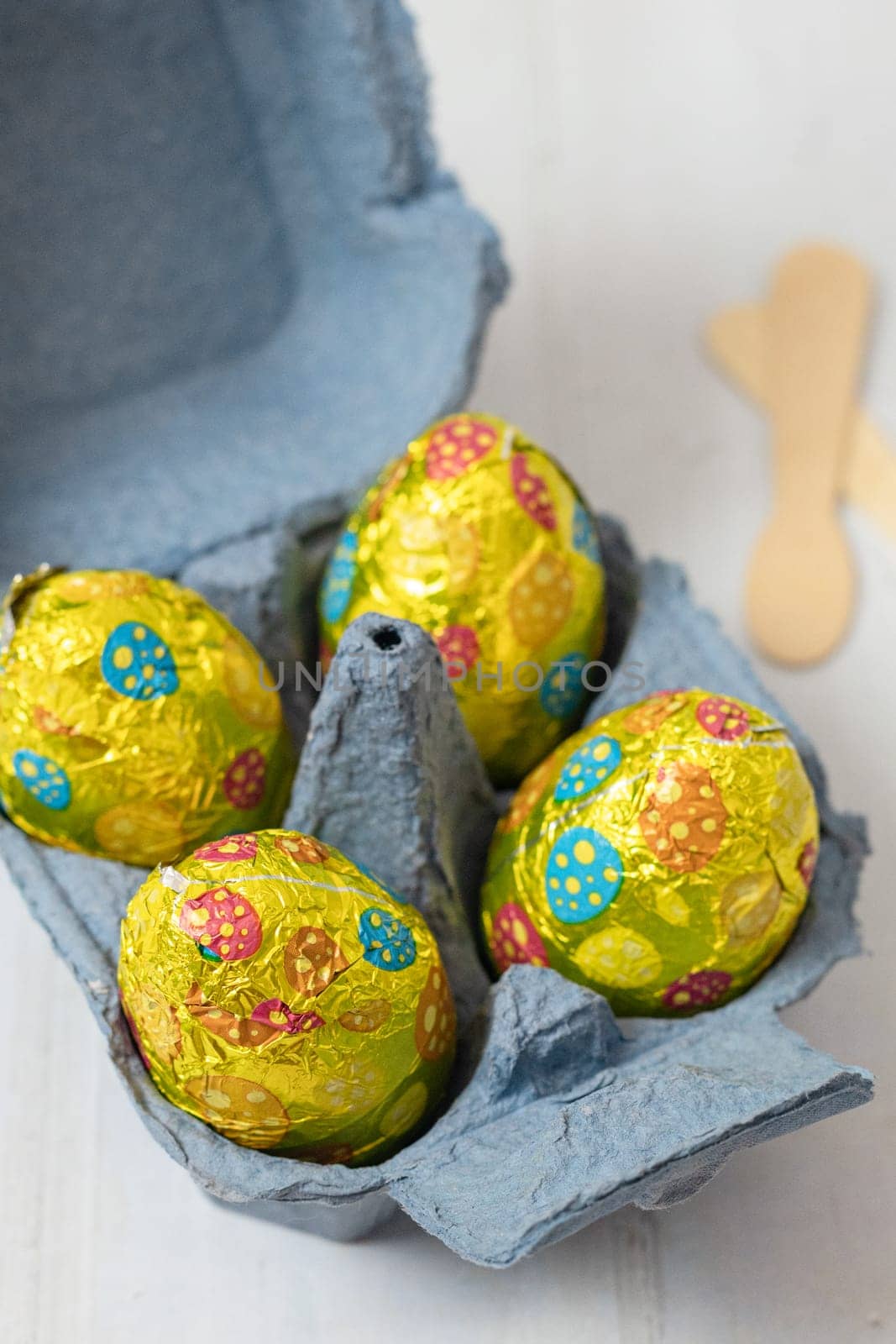 Four Easter eggs in shiny colorful wrappers in a blue egg carton box and two wooden spoons lie on a white wooden table, close-up side view.