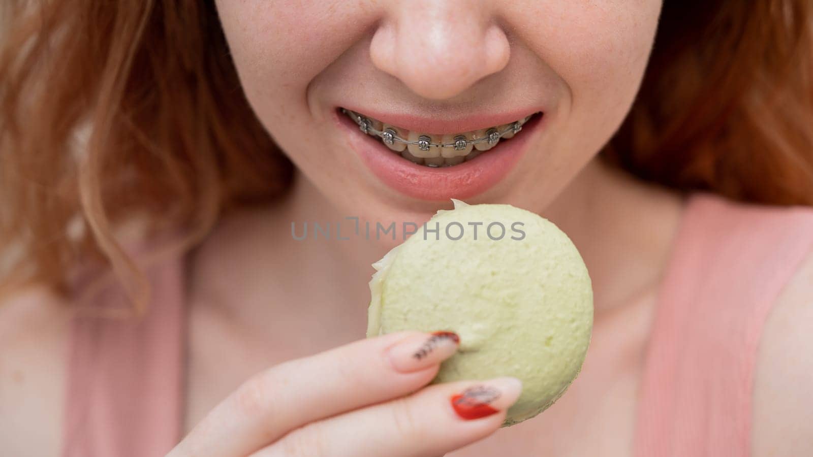 Young red-haired woman with braces eating macaron cake. Cropped