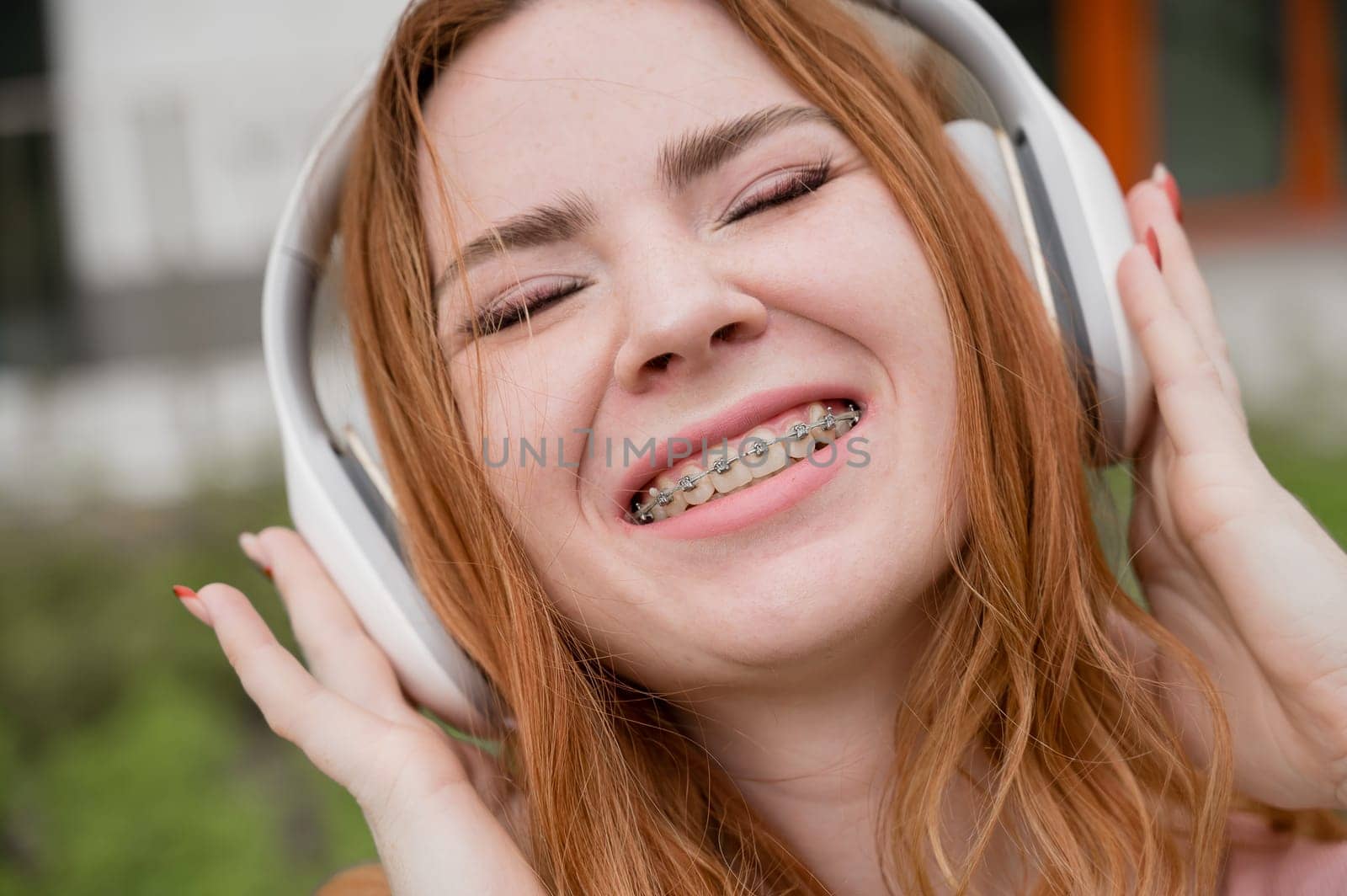 Portrait of a young red-haired woman with braces on her teeth listening to music on headphones outdoors. by mrwed54