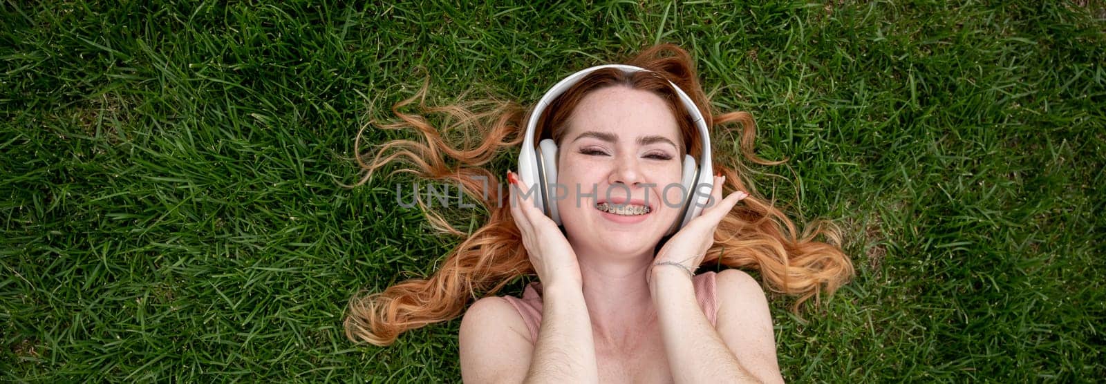 Top view of a young red-haired woman lying on the grass and listening to music on headphones. Widescreen. by mrwed54