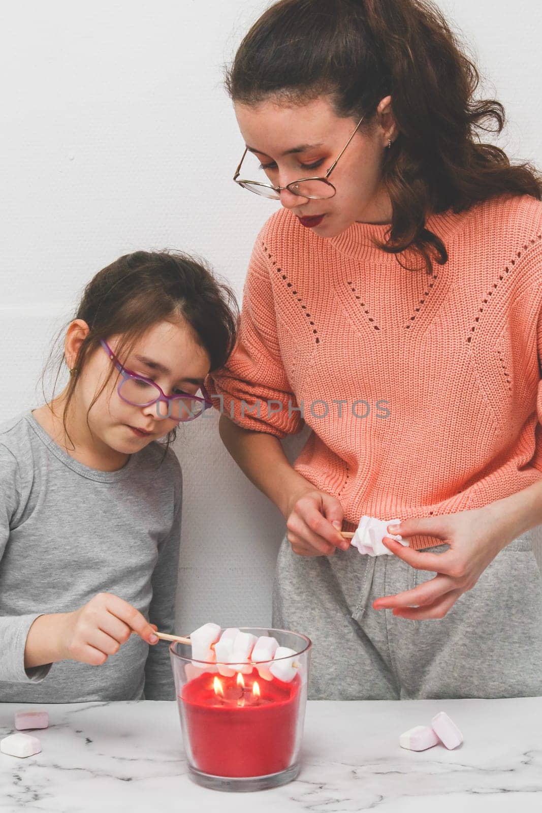 Two little girls in home clothes with disheveled hair with passionate emotion fry marshmallows on a candle fire, holding them on wooden skewers, standing at a light marble table, close-up side view.