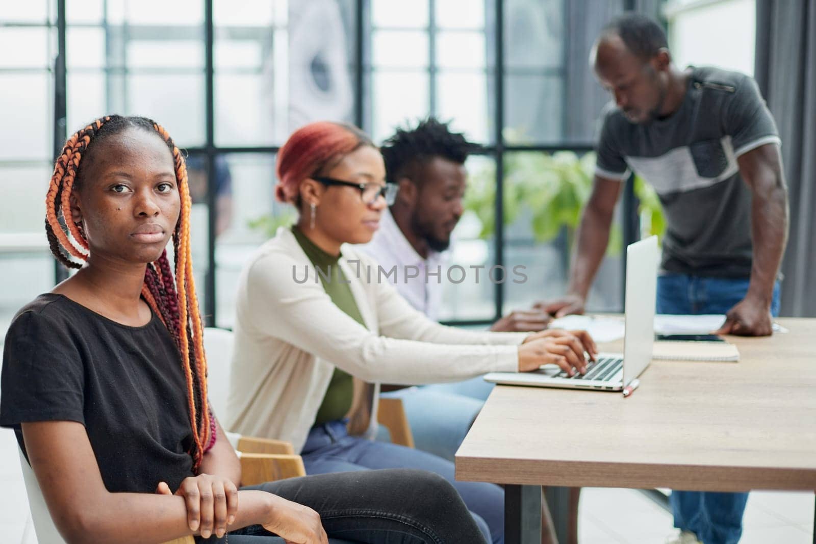 young African American woman in casual clothes posing for the camera while sitting at the table against the background of her colleagues by Prosto