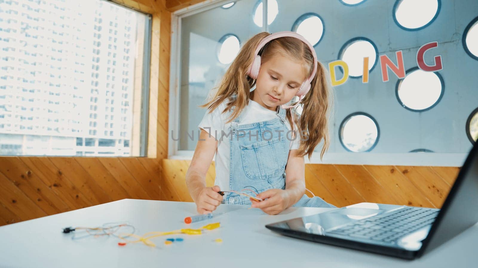 Pretty girl wearing headphone while study electronic equipment. Caucasian child doing science experiment while laptop, screwdriver and wires placed near on table. Smart online classroom. Erudition.