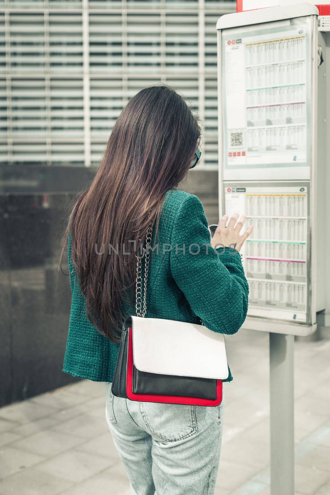 One young caucasian brunette girl with long flowing hair in a green jacket stands with her back at a bus stop near the schedule board, close-up side view.