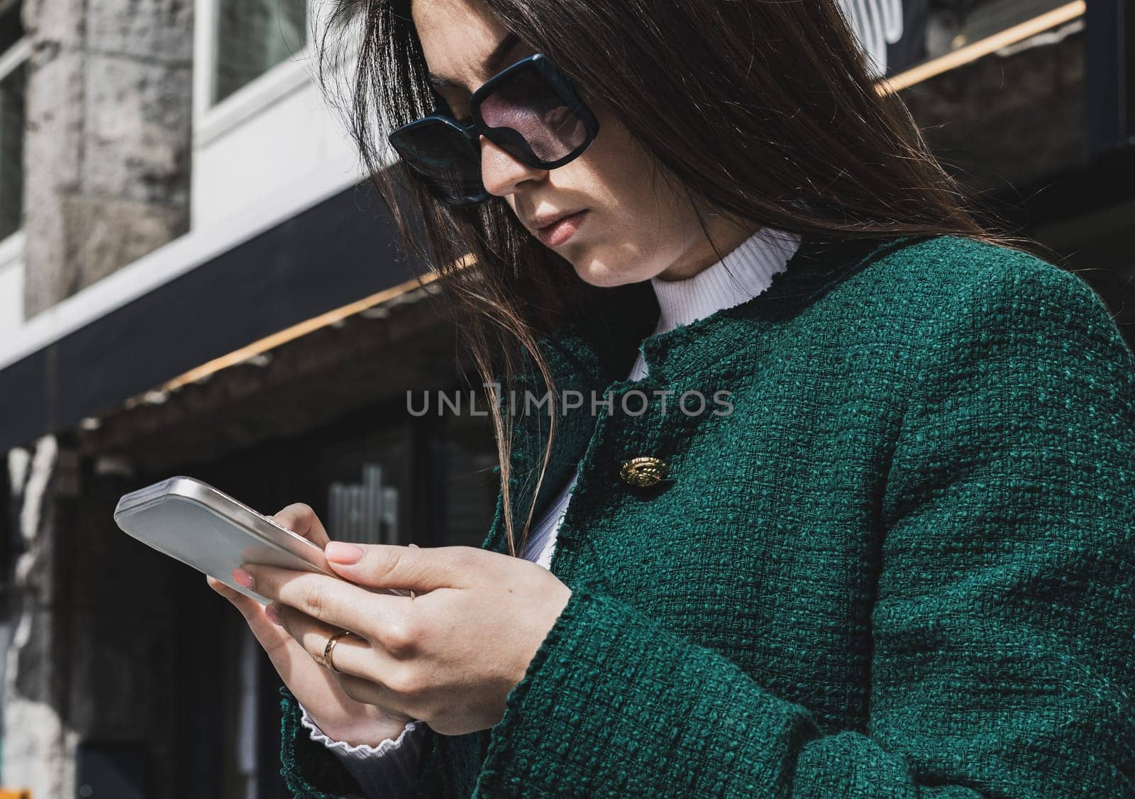 Portrait of one young beautiful caucasian brunette girl with long flowing hair in a green jacket and sunglasses stands on a city street with a mobile phone in her hands, browses the news in social networks, close-up side view.