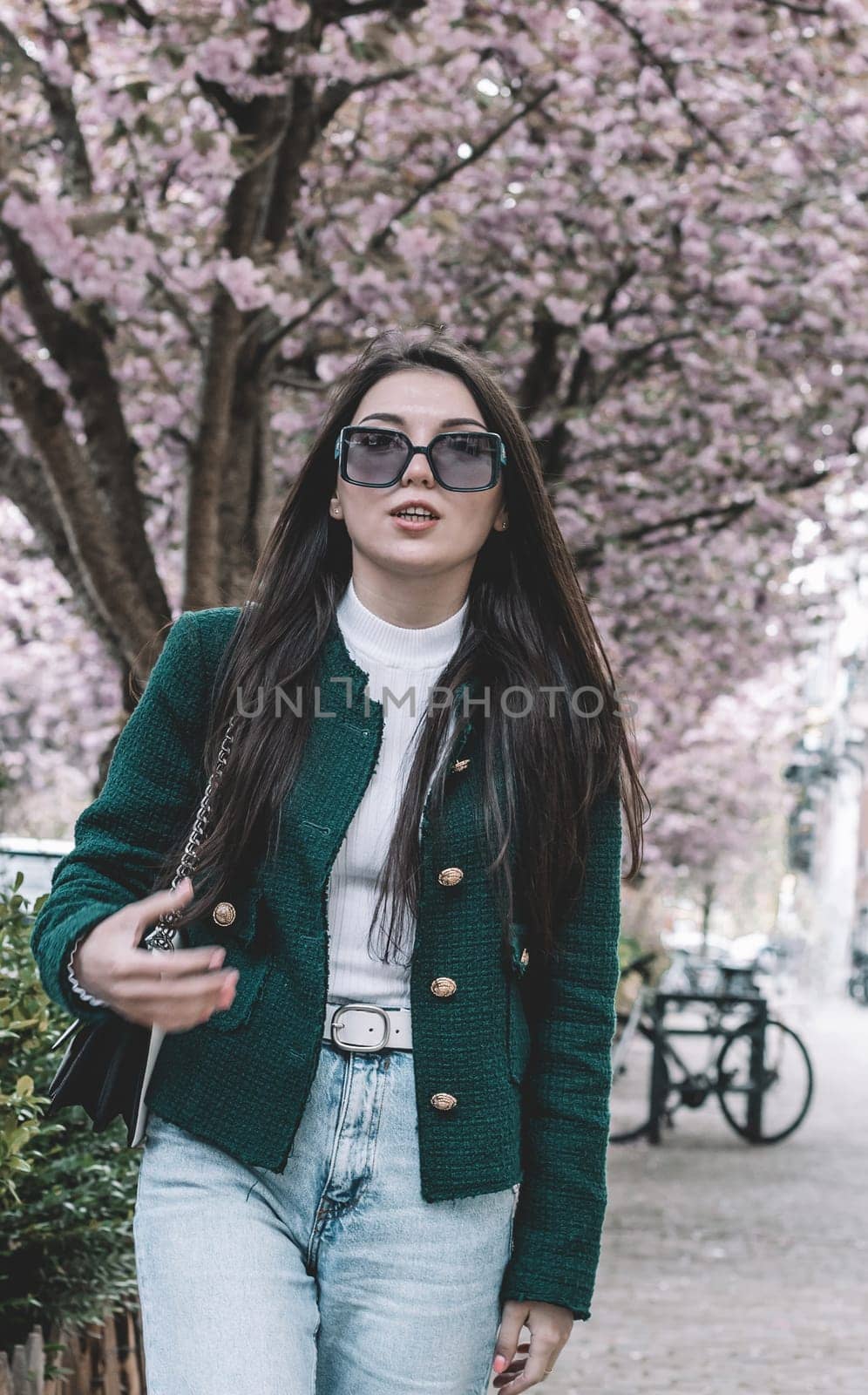 A portrait of one young beautiful Caucasian brunette with long flowing hair in a green jacket and sunglasses, emotionally speaking, walks along a city street with blooming sakura trees on a spring day, close-up view from below.