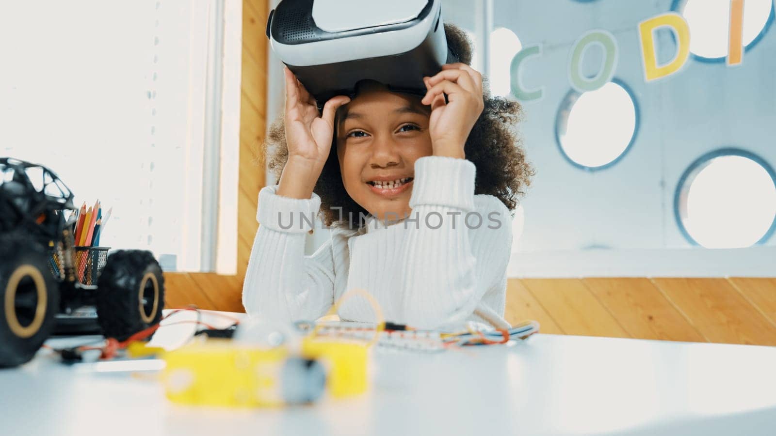 African girl taking off VR glass or head set while looking at camera. Student smiling at camera with colored pencil and laptop placed on table in STEM technology class. Online education. Erudition.