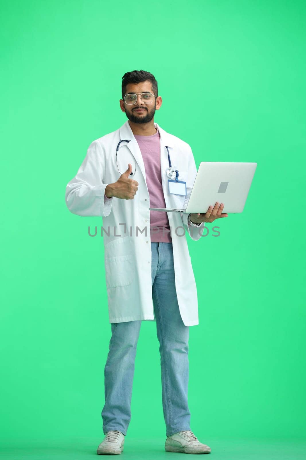 A male doctor, on a green background, in full height, uses a laptop.