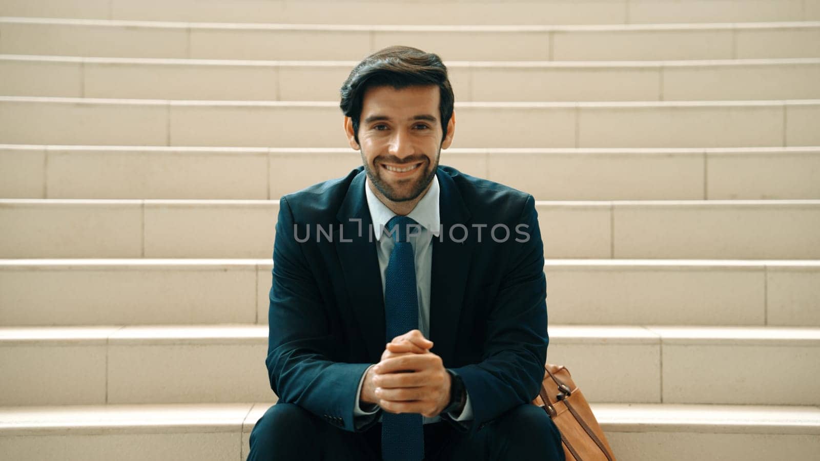 Smiling skilled businessman looking at camera while sitting on stairs. Exultant. by biancoblue