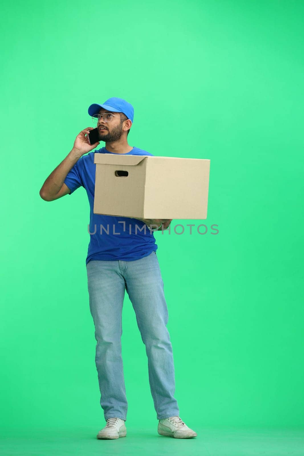 A male deliveryman, on a green background, in full height, with a box and a phone.