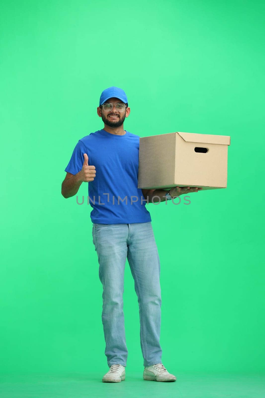 A male deliveryman, on a green background, in full height, with a box, shows a thumbs up.