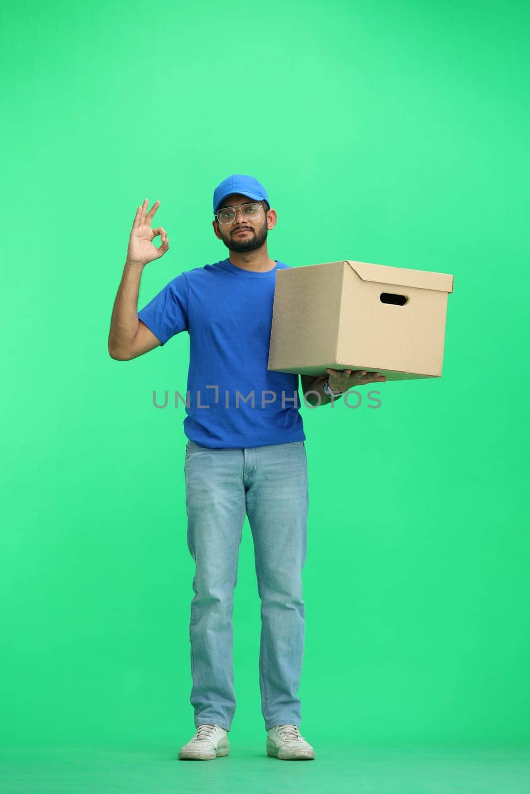 A male deliveryman, on a green background, in full height, with a box, shows the ok sign by Prosto