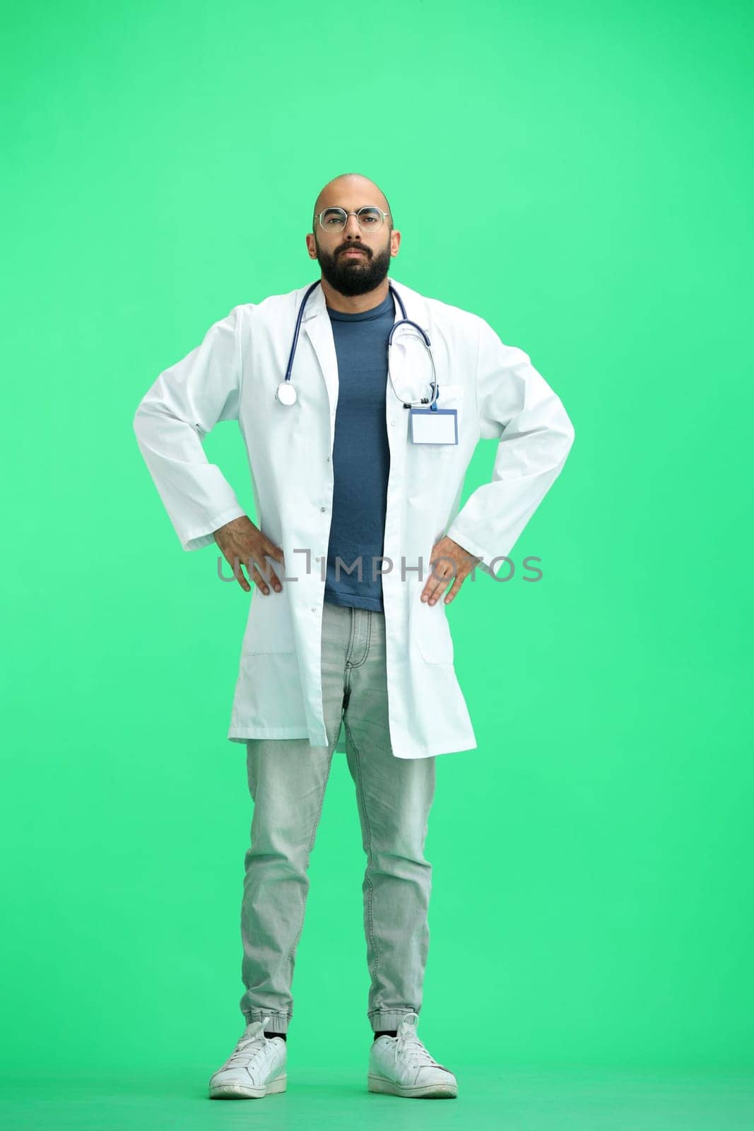 Male doctor, on a green background, full-length, hands on hips.