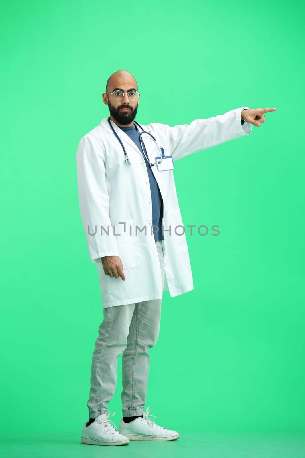 The male doctor, on a green background, in full height, points to the side.