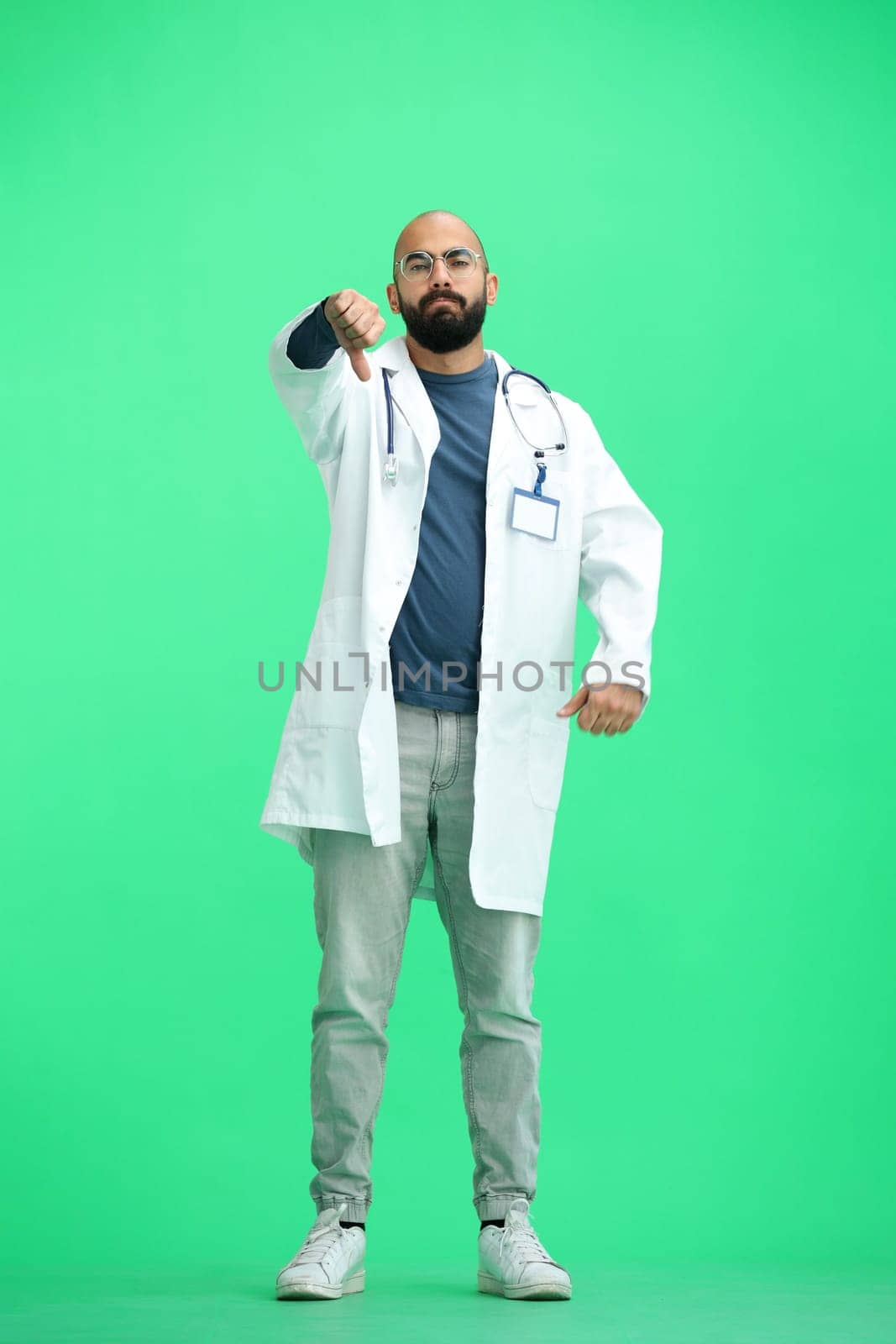 A male doctor, on a green background, in full height, shows his thumbs down.