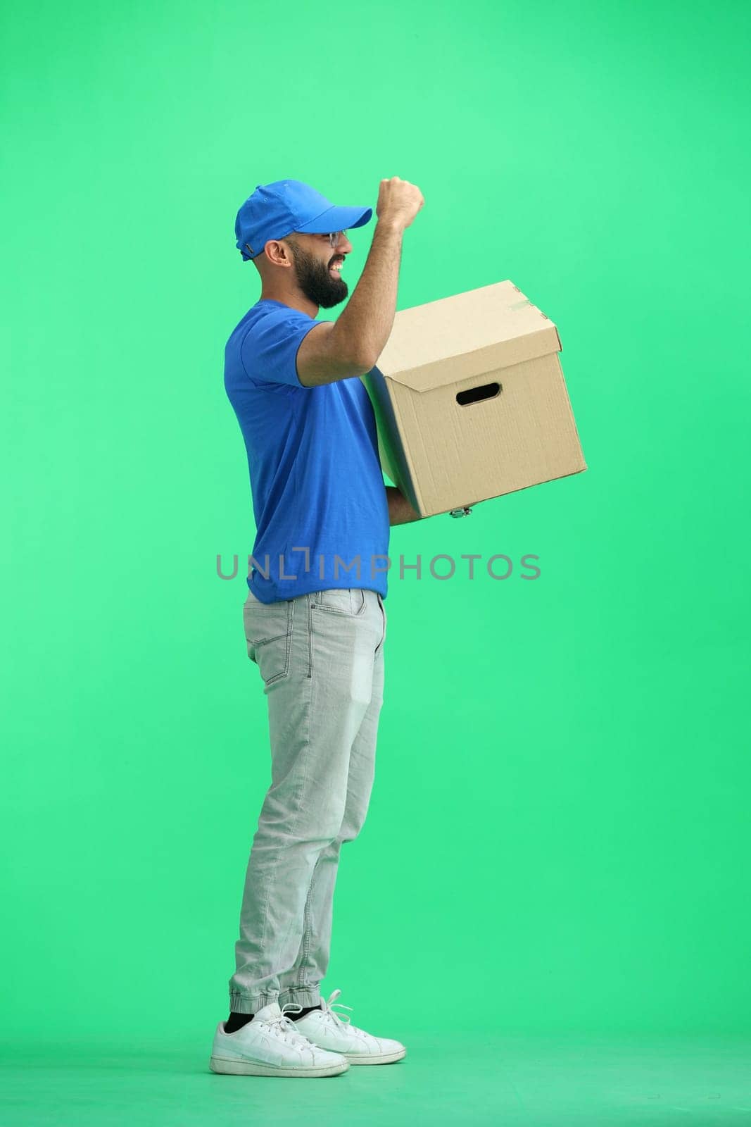 A male deliveryman, on a green background, full-length, with a box, raised his hand up.
