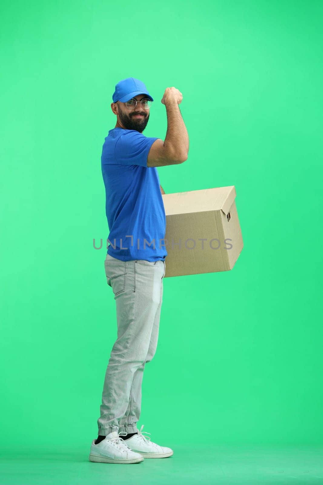 A male deliveryman, on a green background, in full height, with a box, shows strength.