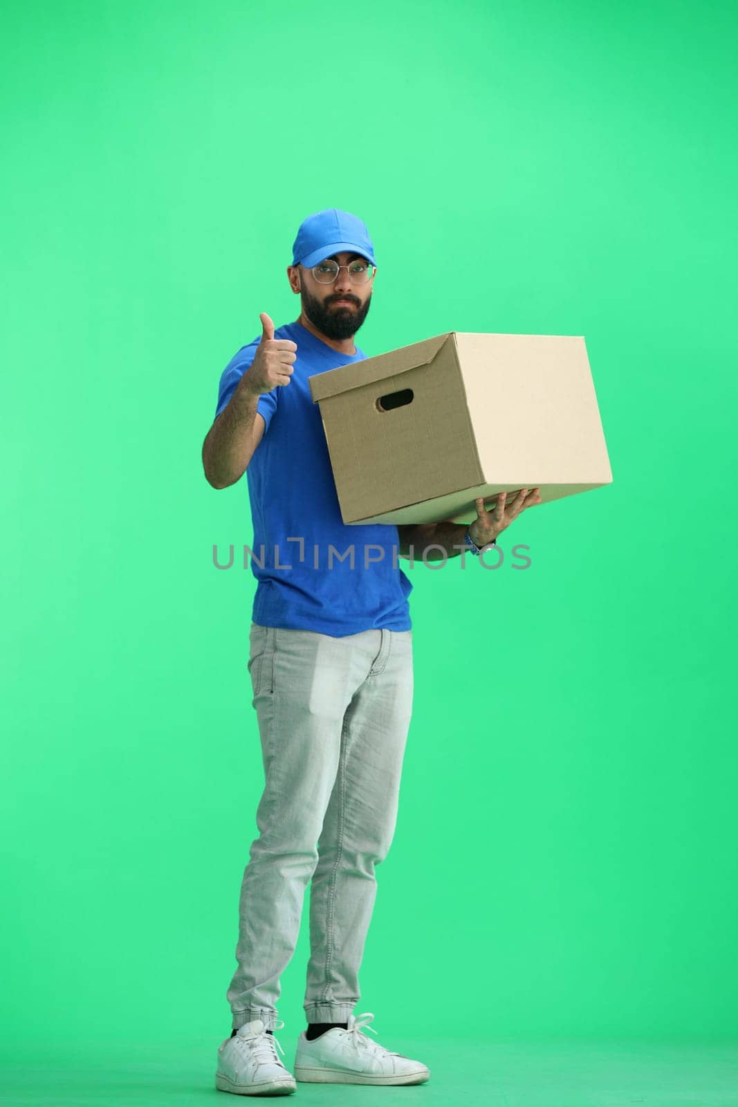 A male deliveryman, on a green background, in full height, with a box, shows a thumbs up.