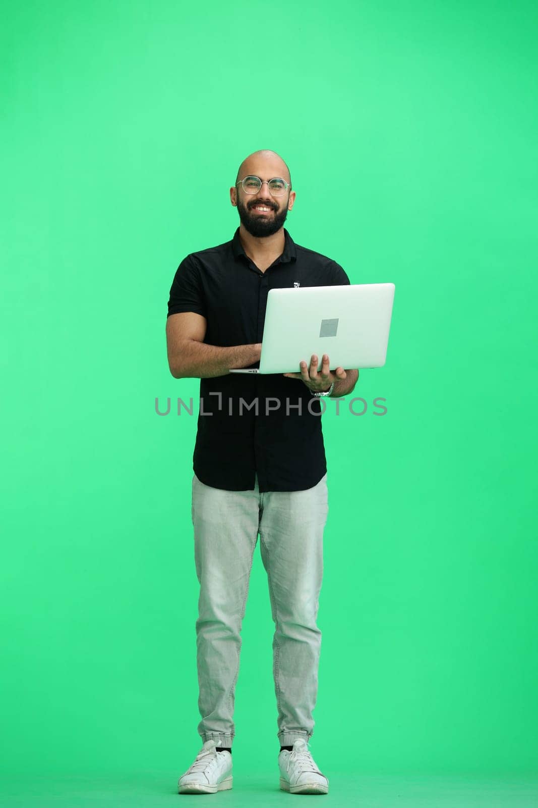 A man, on a green background, in full height, uses a laptop.