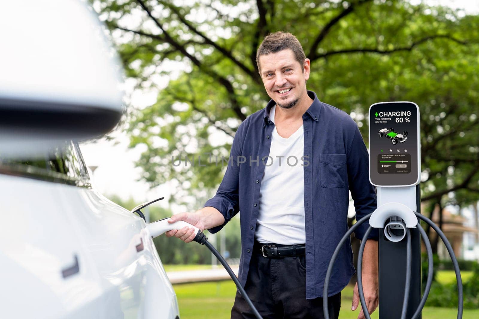 Man recharge EV electric vehicle's battery from EV charging station in outdoor green city park scenic. Eco friendly urban transport and commute with eco friendly EV car travel. Exalt