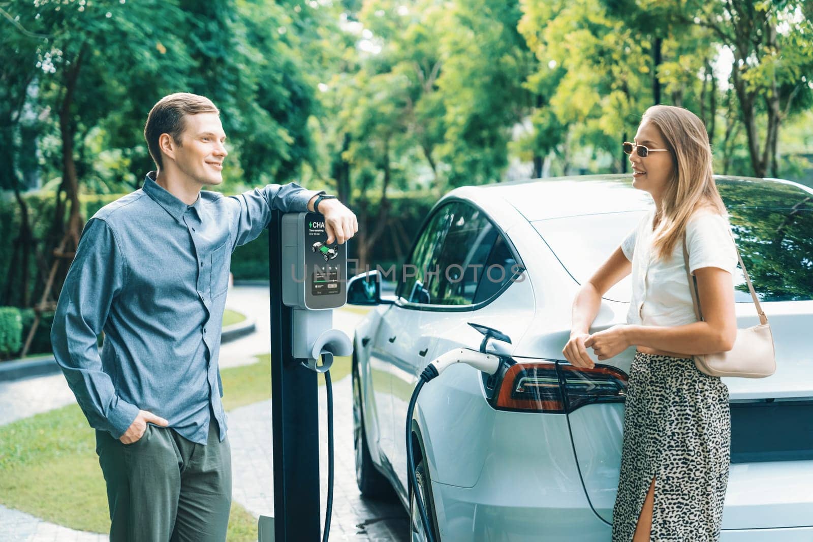 Young couple travel with EV electric car charging in green sustainable city outdoor garden in summer shows urban sustainability lifestyle by green clean rechargeable energy of electric vehicle innards