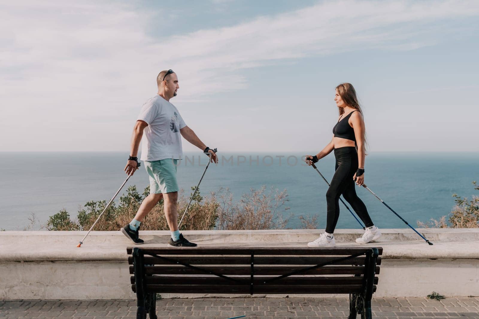 Happy Middle aged couple or friends practicing nordic walking in park near sea. Mature couple with trekking poles walking, practicing Nordic walking outdoors. Aging youthfully and sport concept.