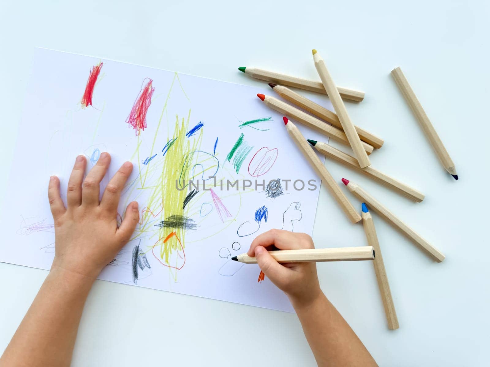 small child draws with colored pencils on paper on white table. by Lunnica