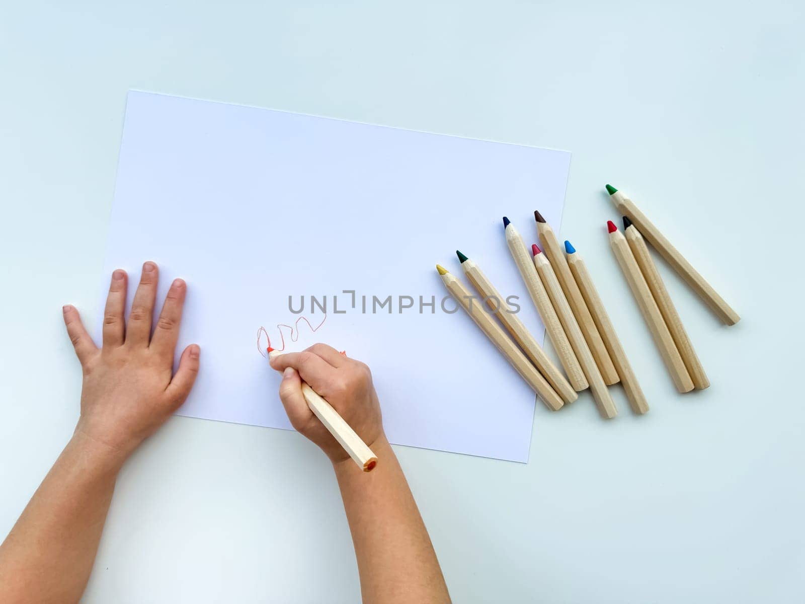 small child draws with colored pencils on paper on white table. by Lunnica