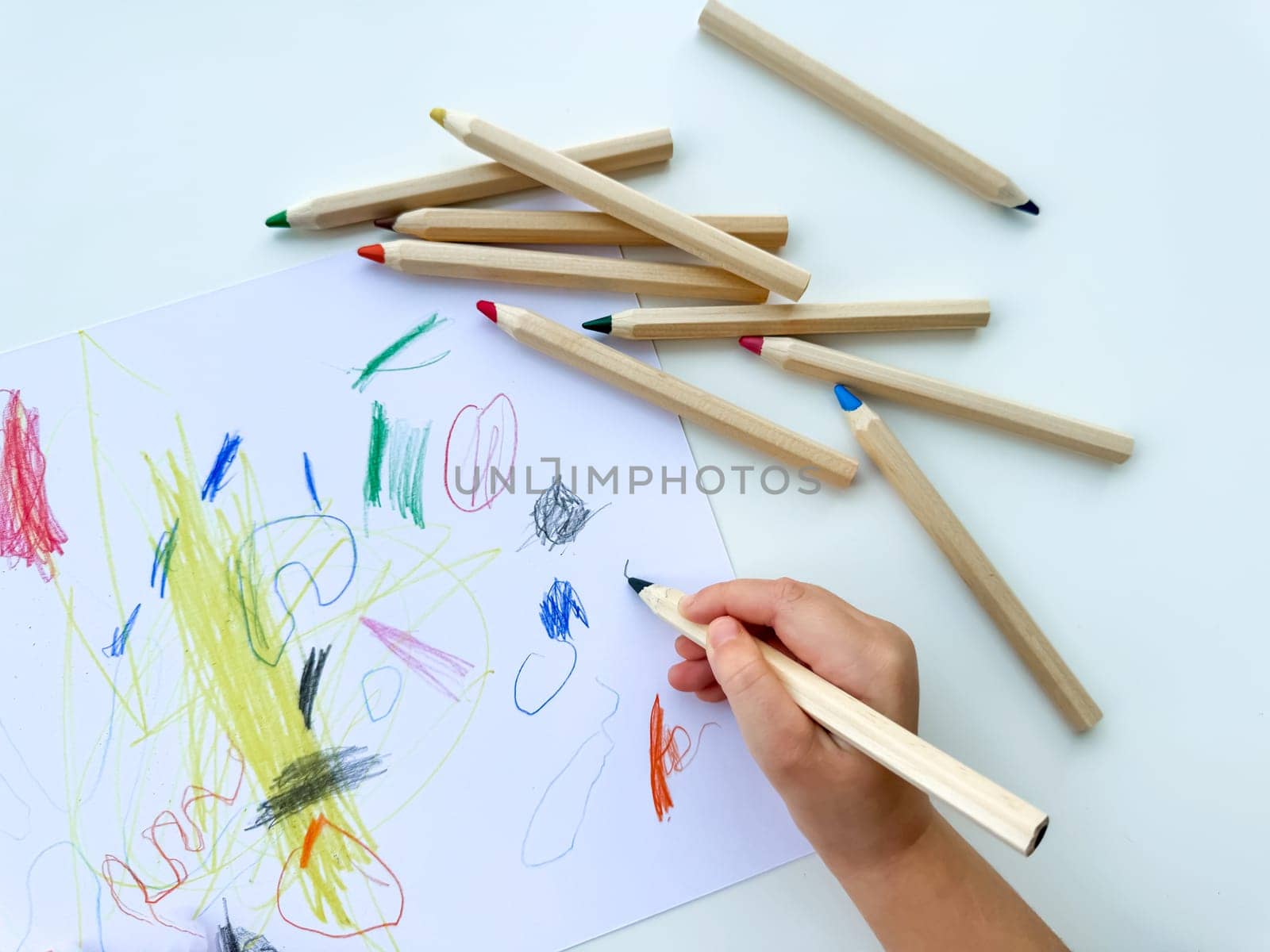 small child draws with colored pencils on paper on white table. High quality photo