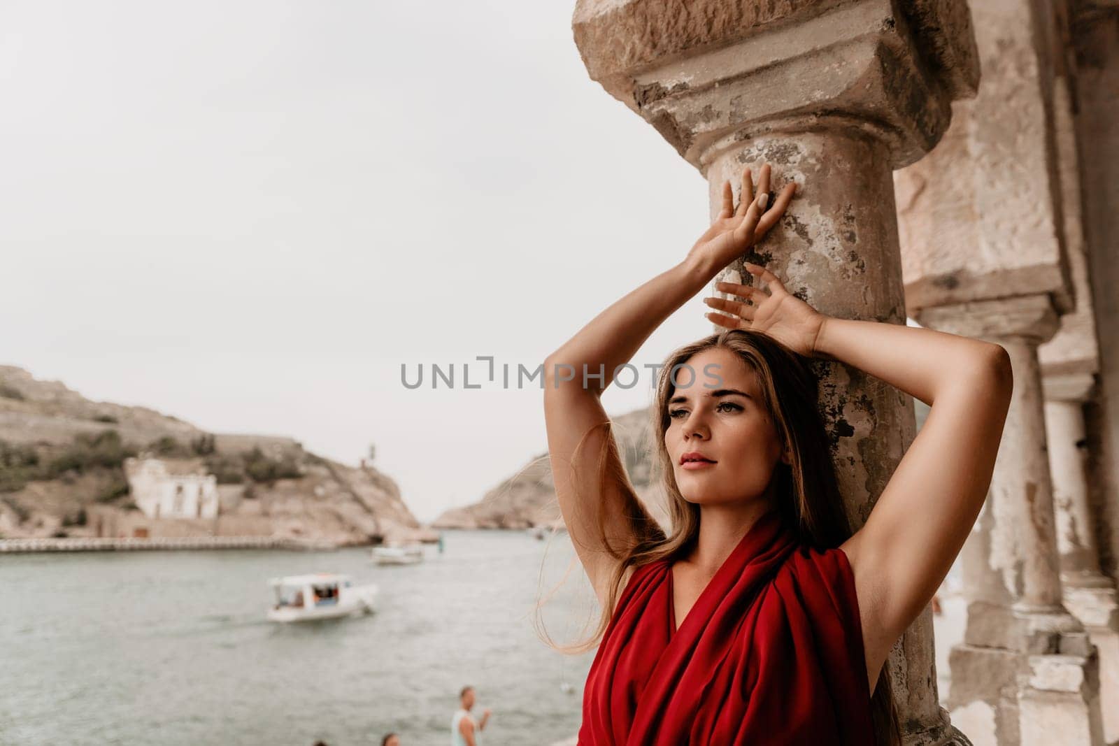 Woman travel portrait. Happy woman with long hair looking at camera and smiling. Close up portrait cute woman in a res long dress posing on backdrop of old travel city near sea by panophotograph