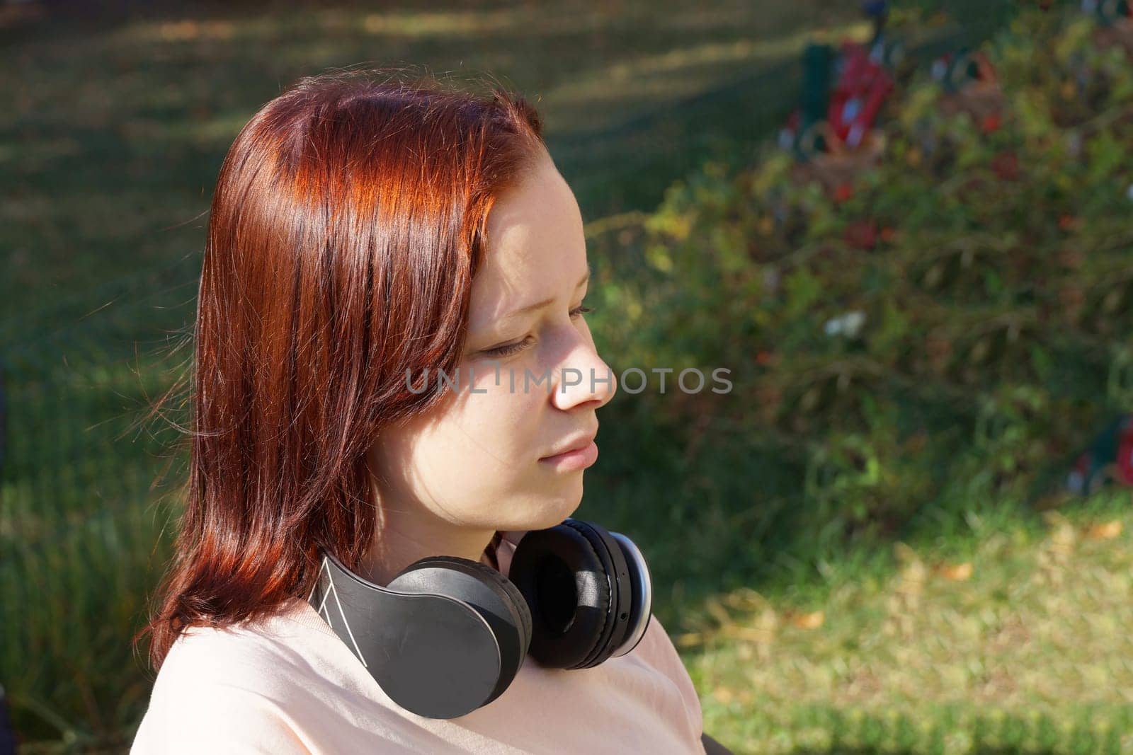 red-haired teenage girl sitting with her eyes closed in headphones in the park by Annado