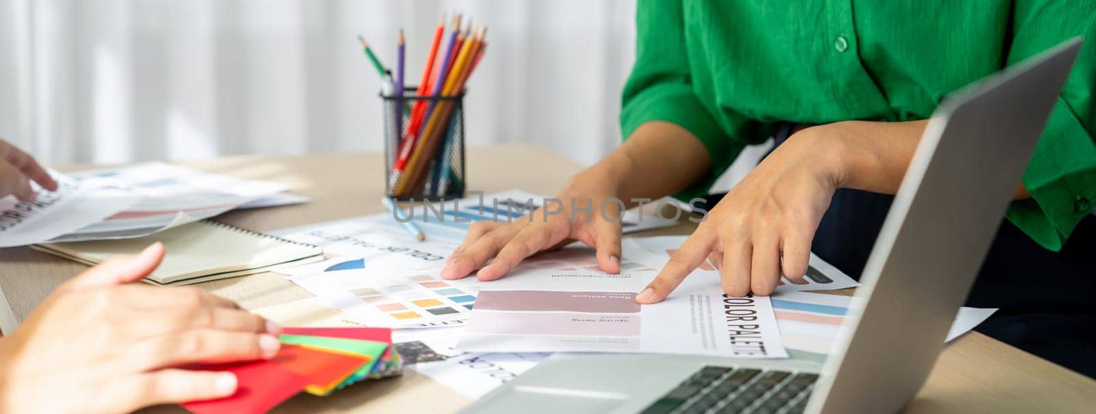 Creative graphic design presents selective color while manager using laptop comparing suitable color at table with color palettes, graphic material scatter around at modern office. Variegated.