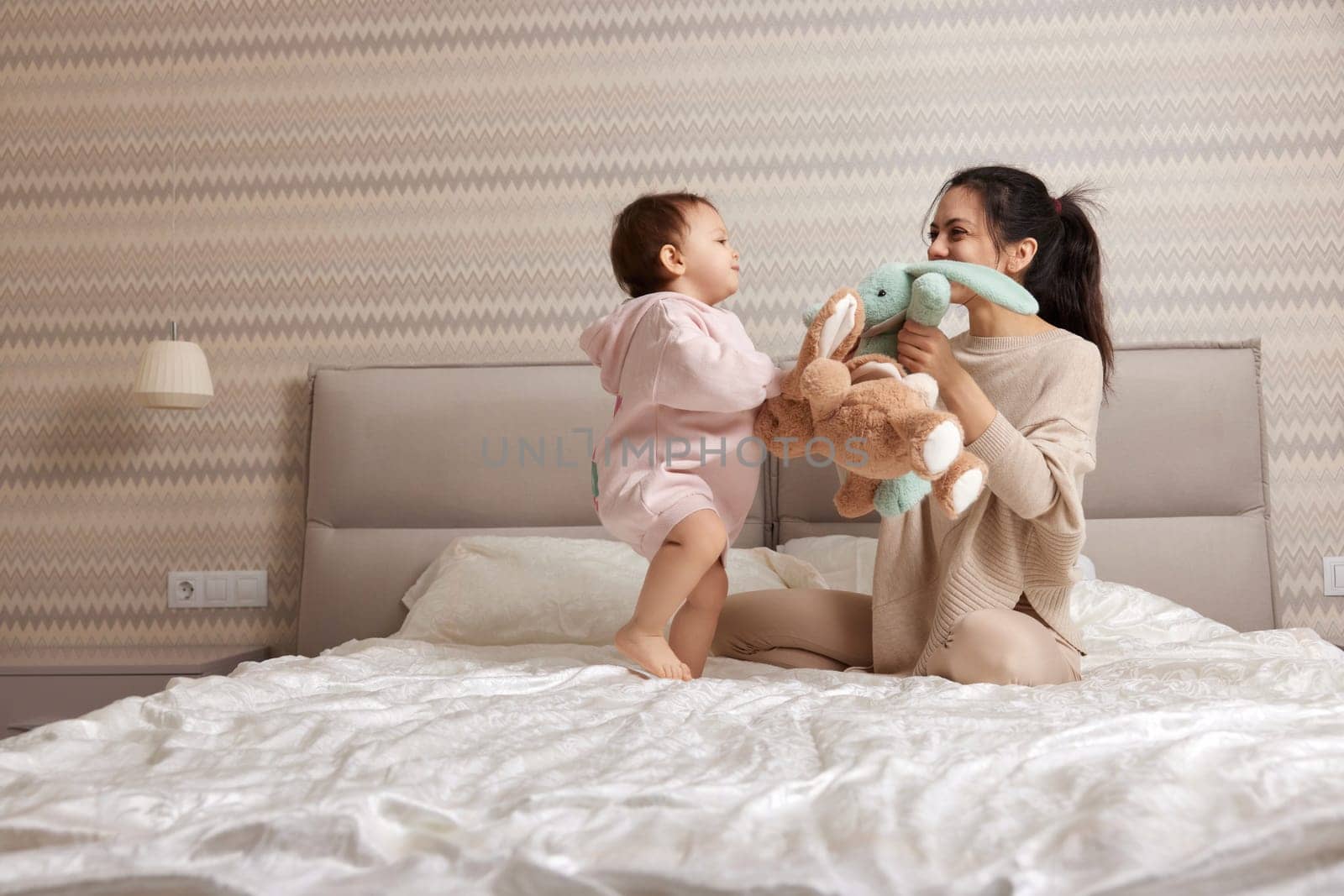 happy mother and her little child daughter playing with bunny toys in bedroom, family having fun