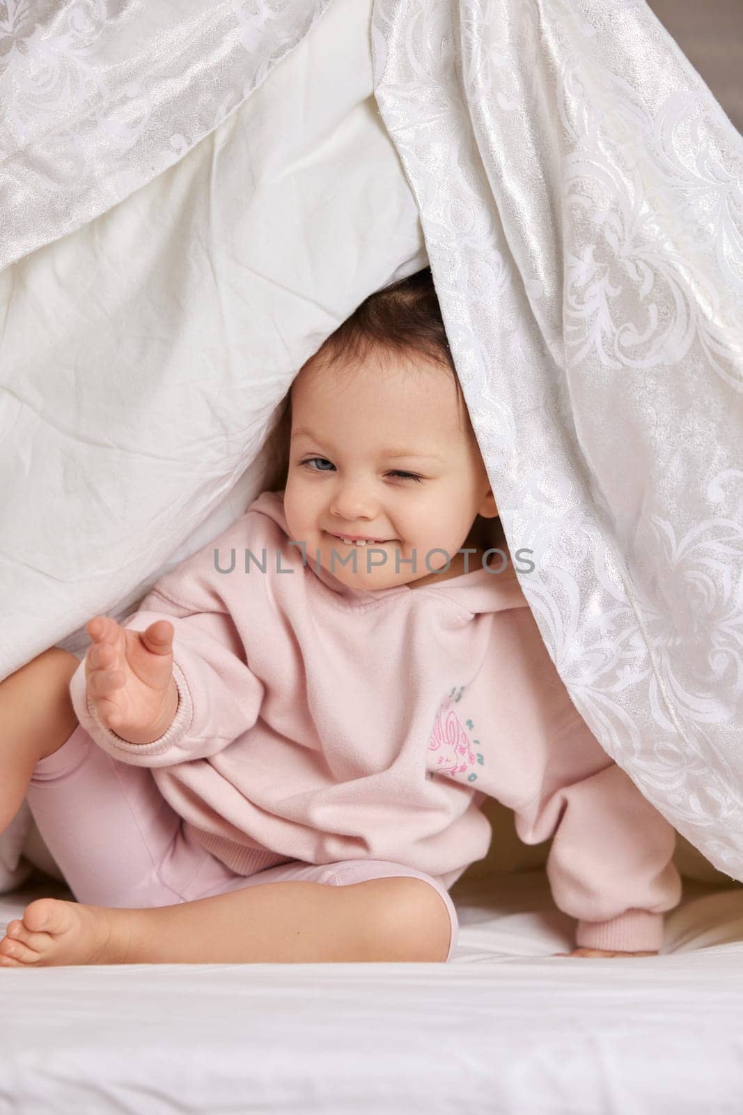 cute funny little child girl looks out from under the blanket on the bed
