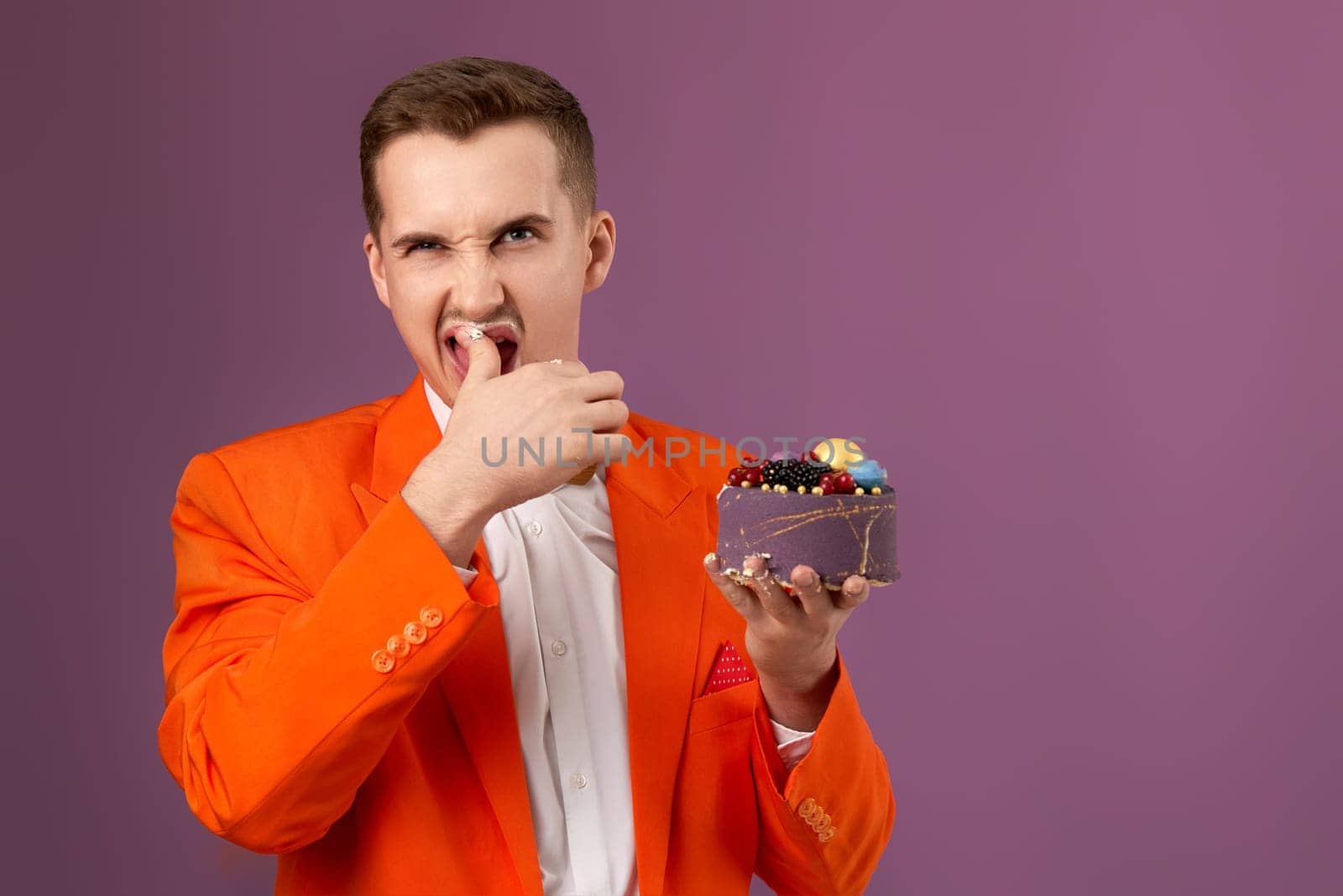 handsome birthday man in orange jacket eating cake on purple background