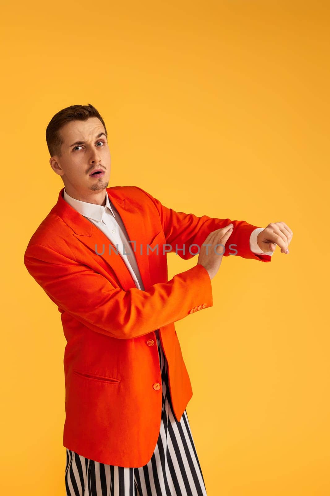 Cheerful funny young man in orange jacket and striped pants on yellow background.