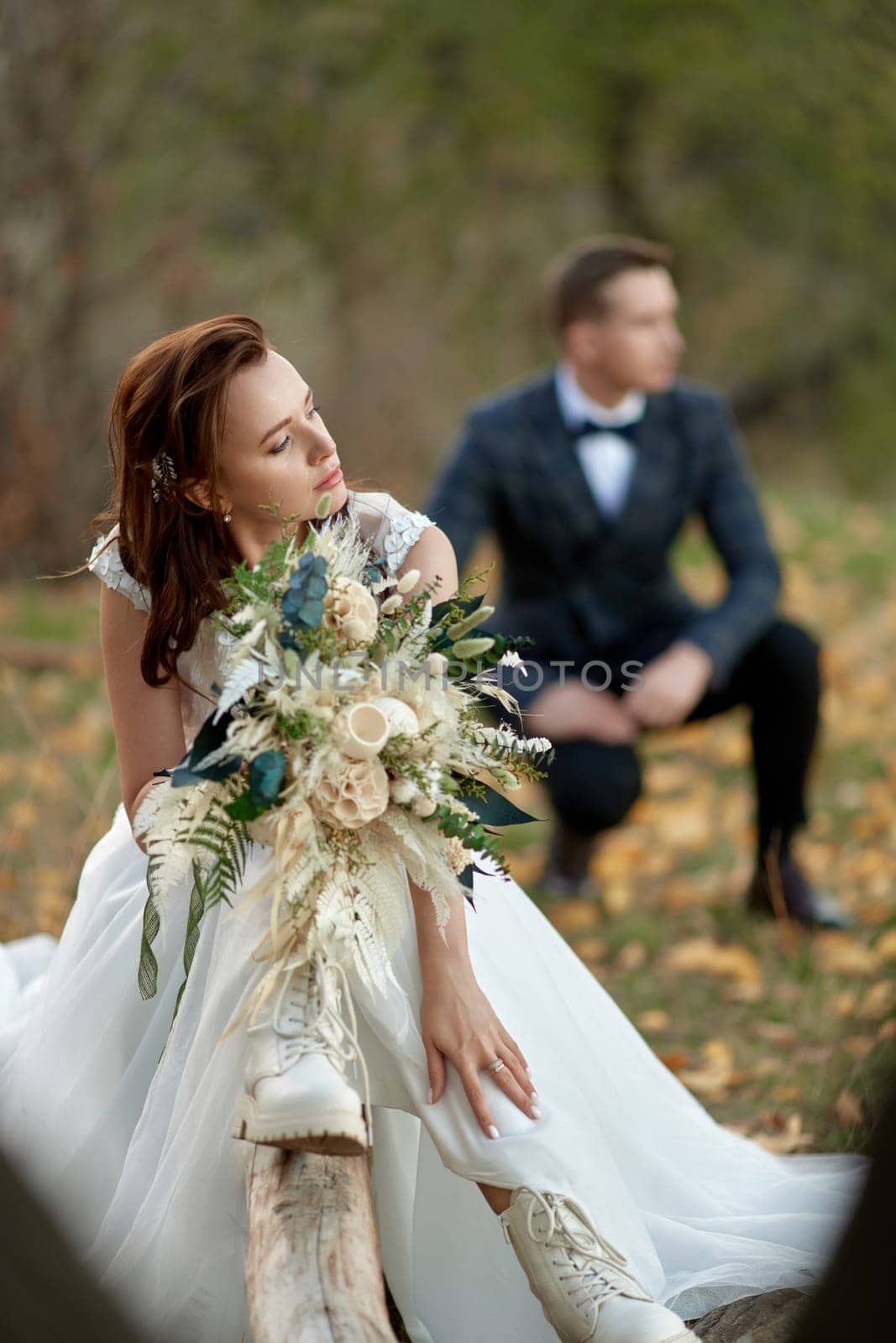 beautiful bride in white boots sitting on tree and holding autumn bouquet. blur groom on the background